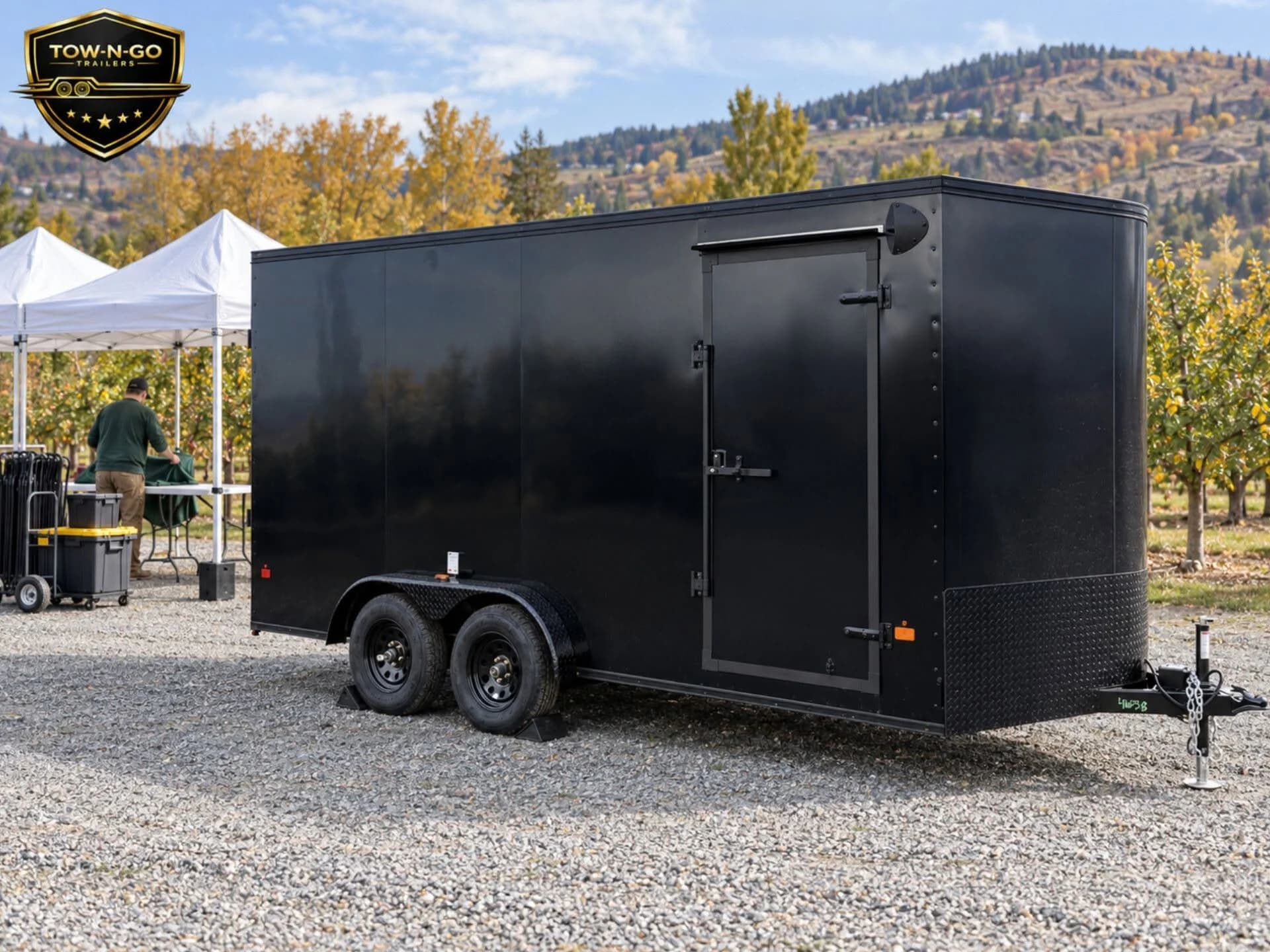 Black enclosed trailer beside white event tents, chairs and equipment cases in an autumn orchard.